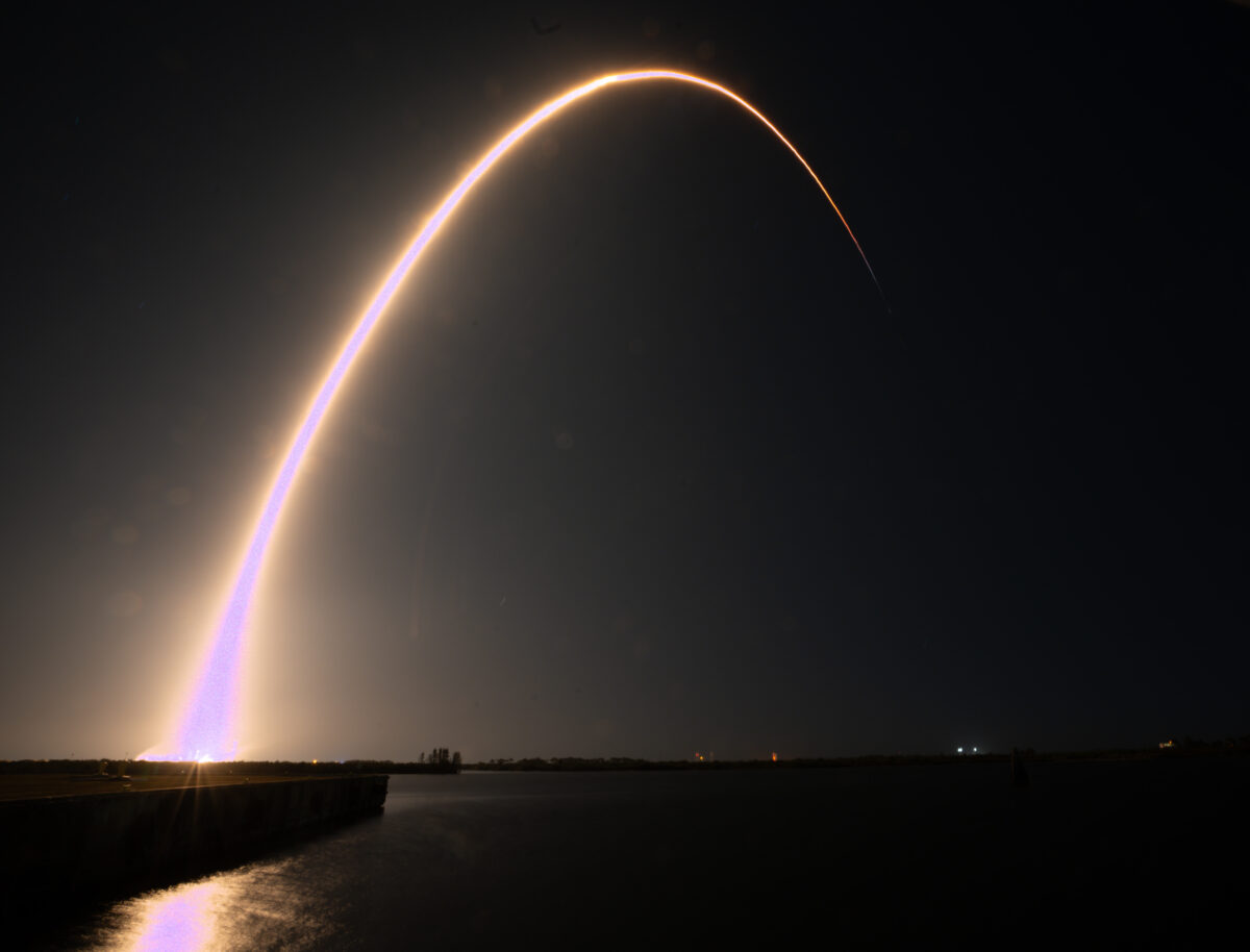 A beam of light extends curved into the sky in a long-exposure photo of a SpaceX launch.