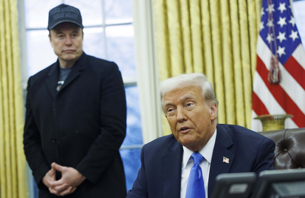 Donald Trump sits at his desk in the oval office, and Elon Musk stands behind him looking at him as he speaks. An American flag stands behind the two of them.