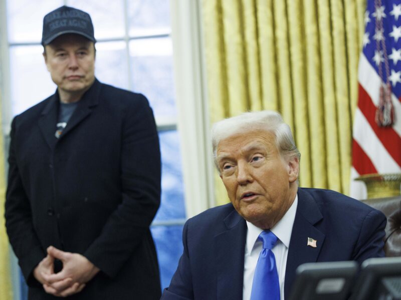 Donald Trump sits at his desk in the oval office, and Elon Musk stands behind him looking at him as he speaks. An American flag stands behind the two of them.
