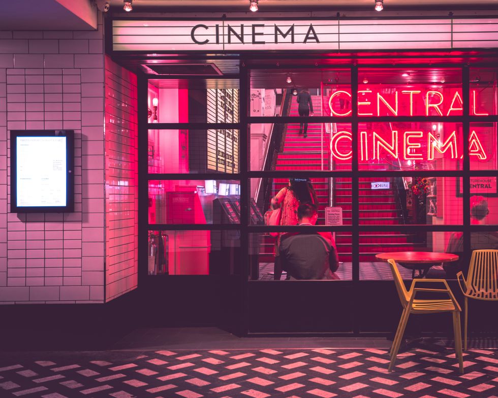 A cinema is seen from the outside. Inside the windows are neon signs, red velvet staircases, and a board showing the movies playing, as well as a handful of people milling about.