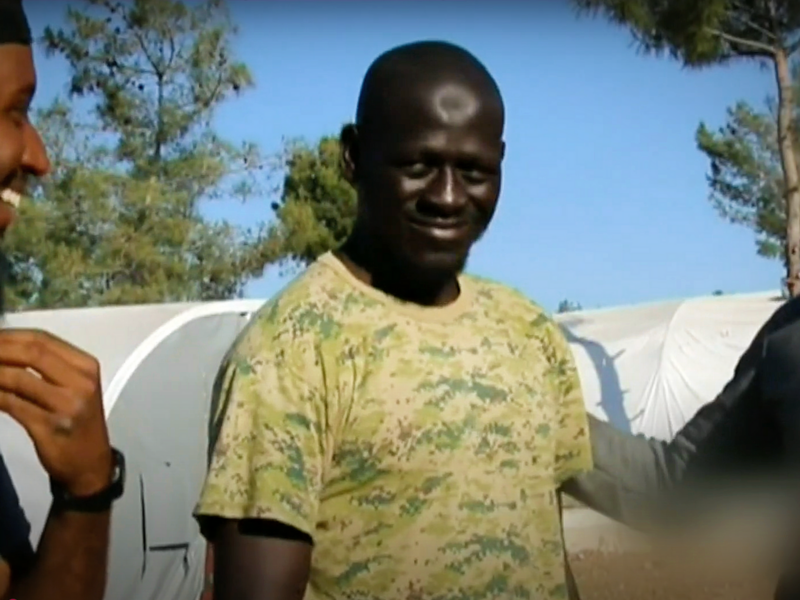 A man wearing a camo shirt stands in front of a series of white tents.