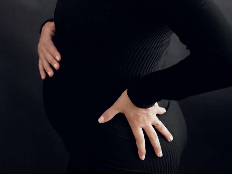 A pregnant woman in a black dress holds her belly with one hand against a black background