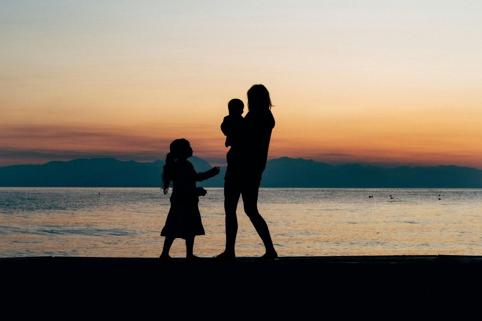 A woman, a young girl, and a baby stand on the beach at dusk, making only their outline visible against the brilliant sky, ocean and mountains in the distance.