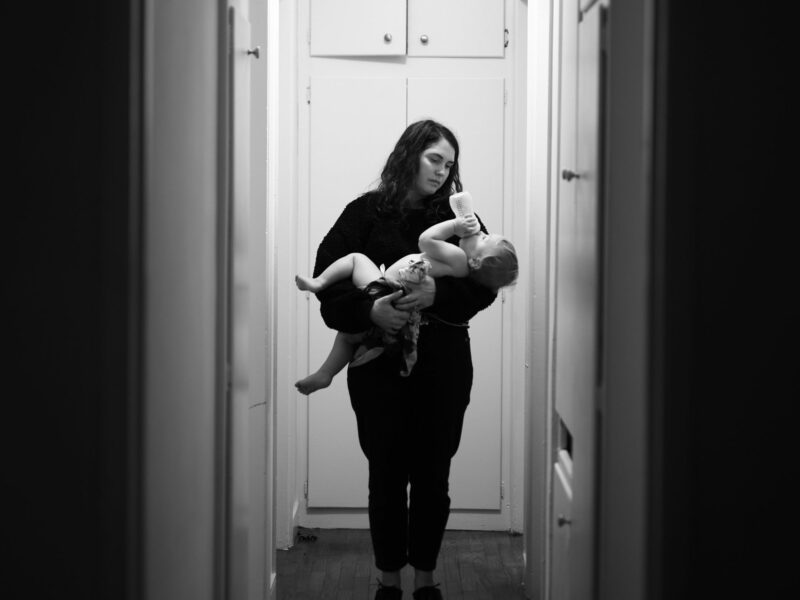A woman holds a baby at the end of a dimly lit corridor. The photo is black and white.