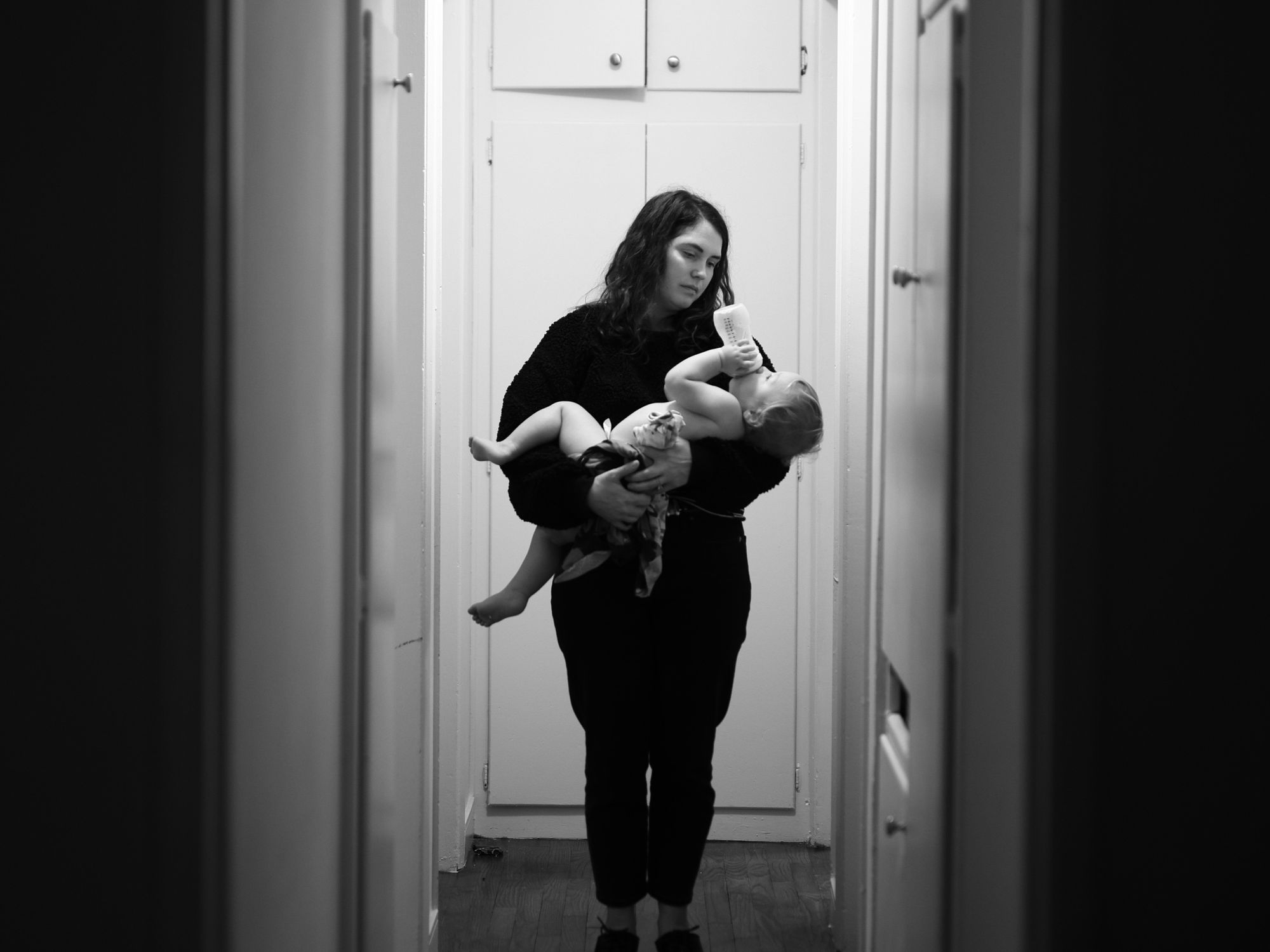 A woman holds a baby at the end of a dimly lit corridor. The photo is black and white.