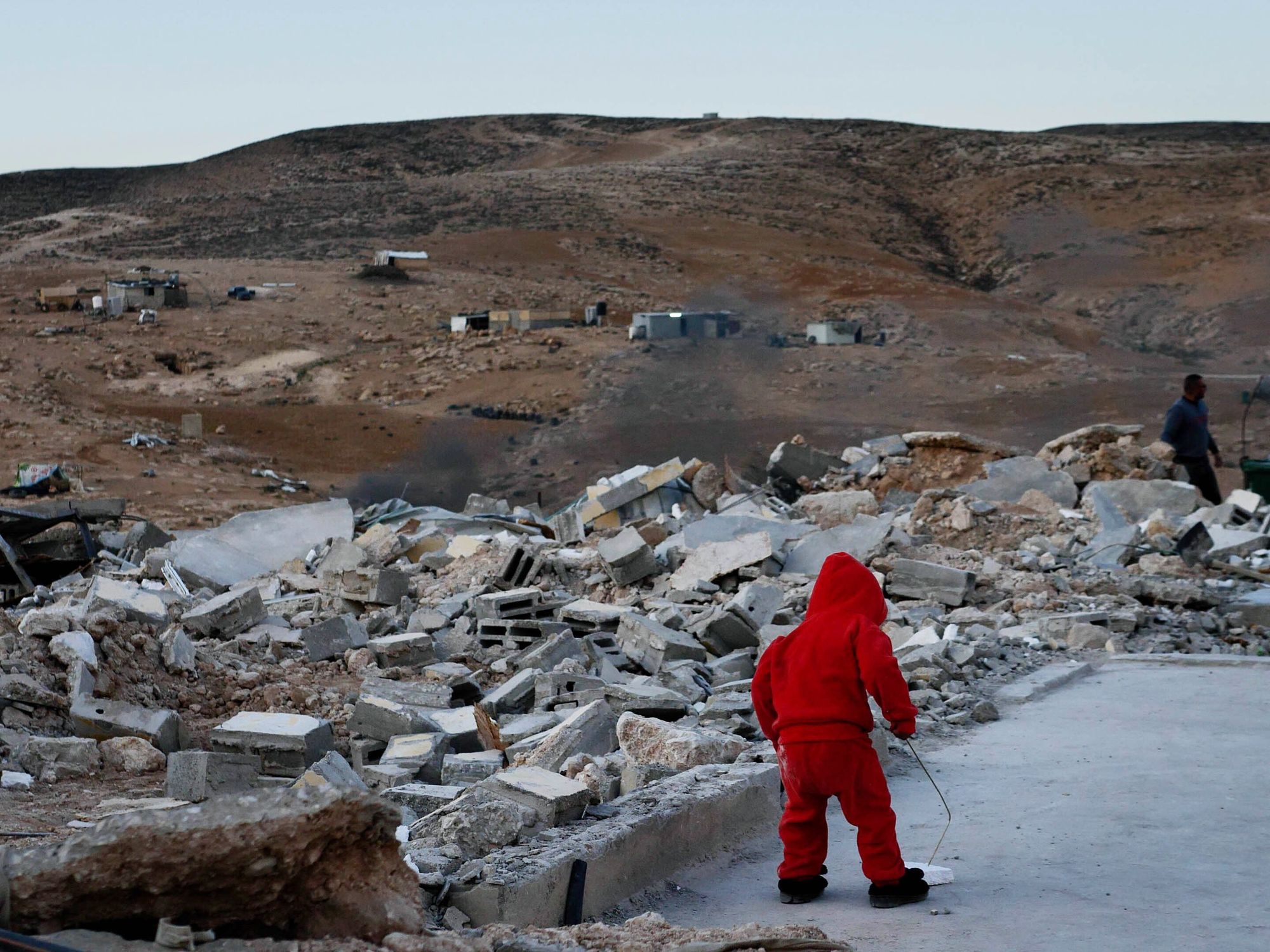 ​A child wearing red stands amidst rubble in a desolate field