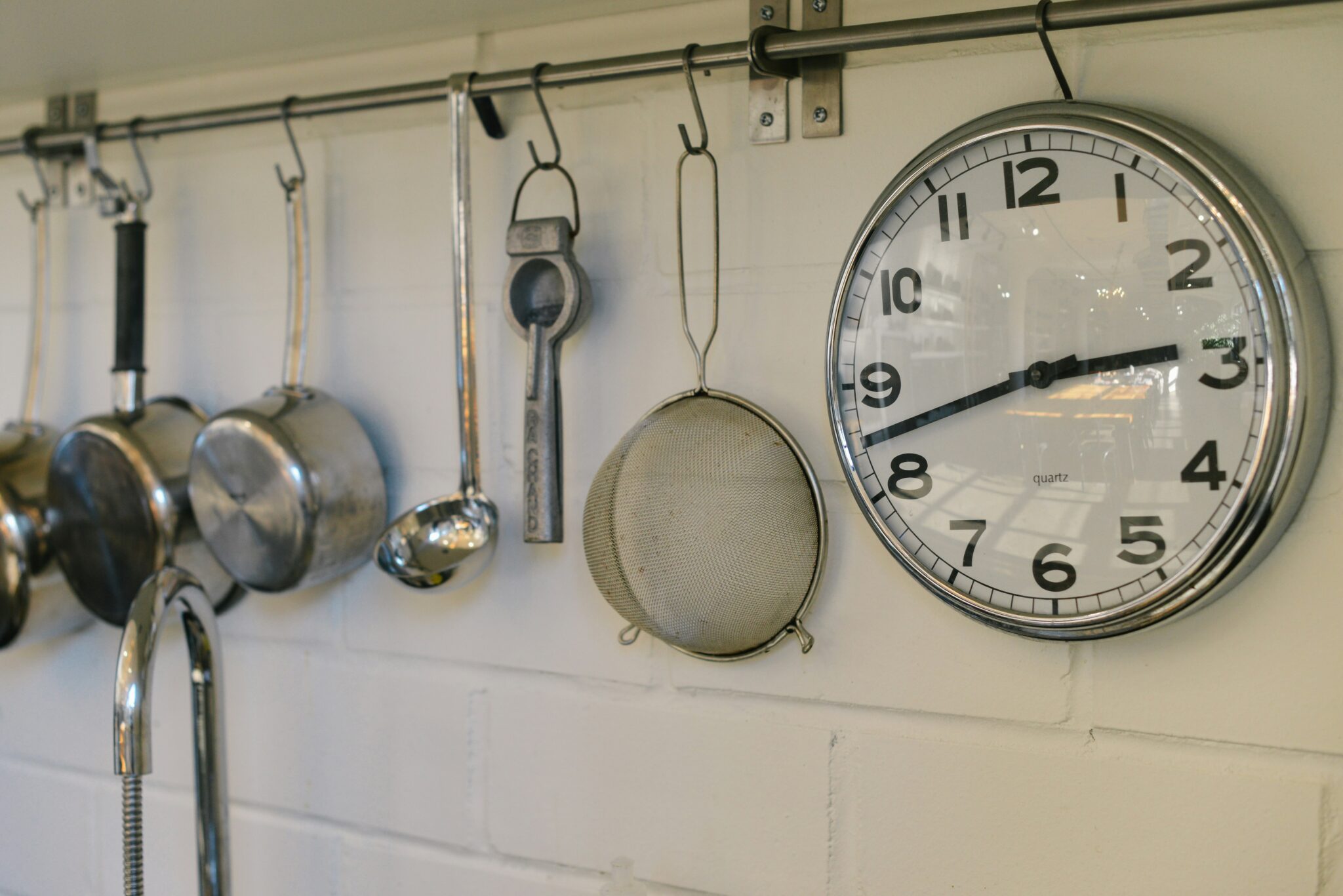 A collander, pots, and other kitchen tools hang alongside a plain white clock.