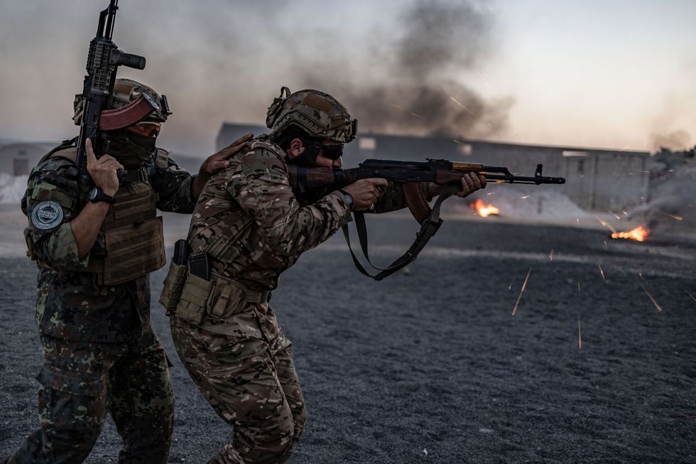 Two men in camo military uniforms crouch together with guns, with flames and smoke around buildings behind them.