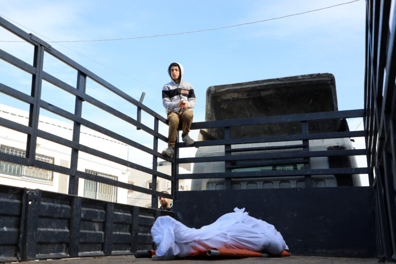 Photo of a man watching over the body of a man draped in a white cloth, lying at the back of a truck. The man was slain in an Israeli raid in central Gaza on Jan. 26.