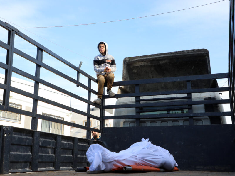 Photo of a man watching over the body of a man draped in a white cloth, lying at the back of a truck. The man was slain in an Israeli raid in central Gaza on Jan. 26.