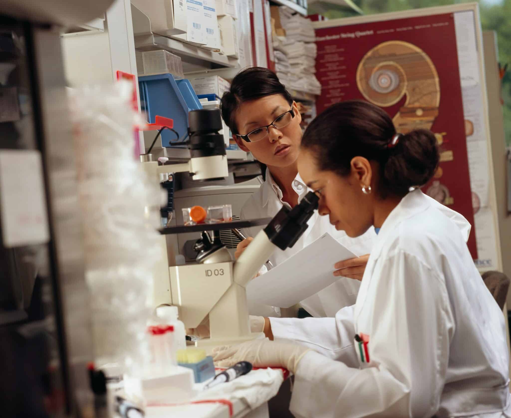 Two women in white lab coats work in a laboratory. One of them looks into a microscope, while the other looks at her from the side.