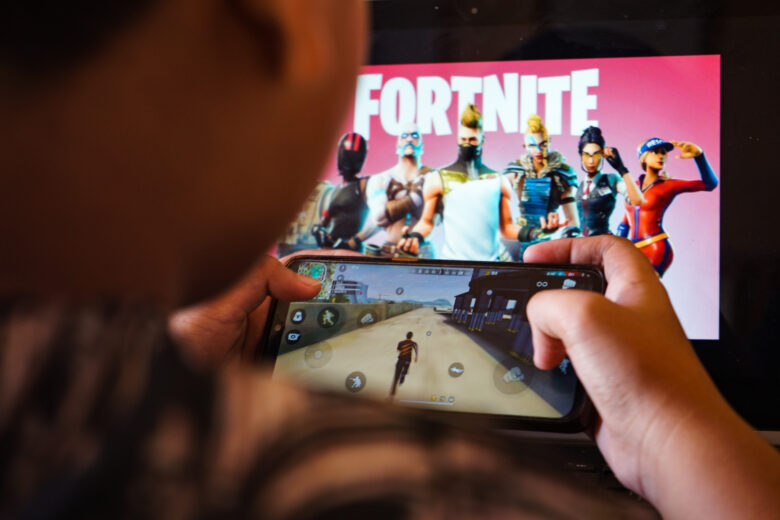 A child plays a game on a cellphone with an image of the Fortnite game on the computer screen in the background.
