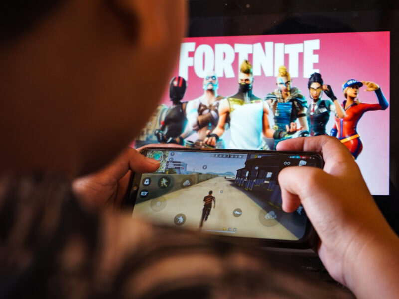 A child plays a game on a cellphone with an image of the Fortnite game on the computer screen in the background.