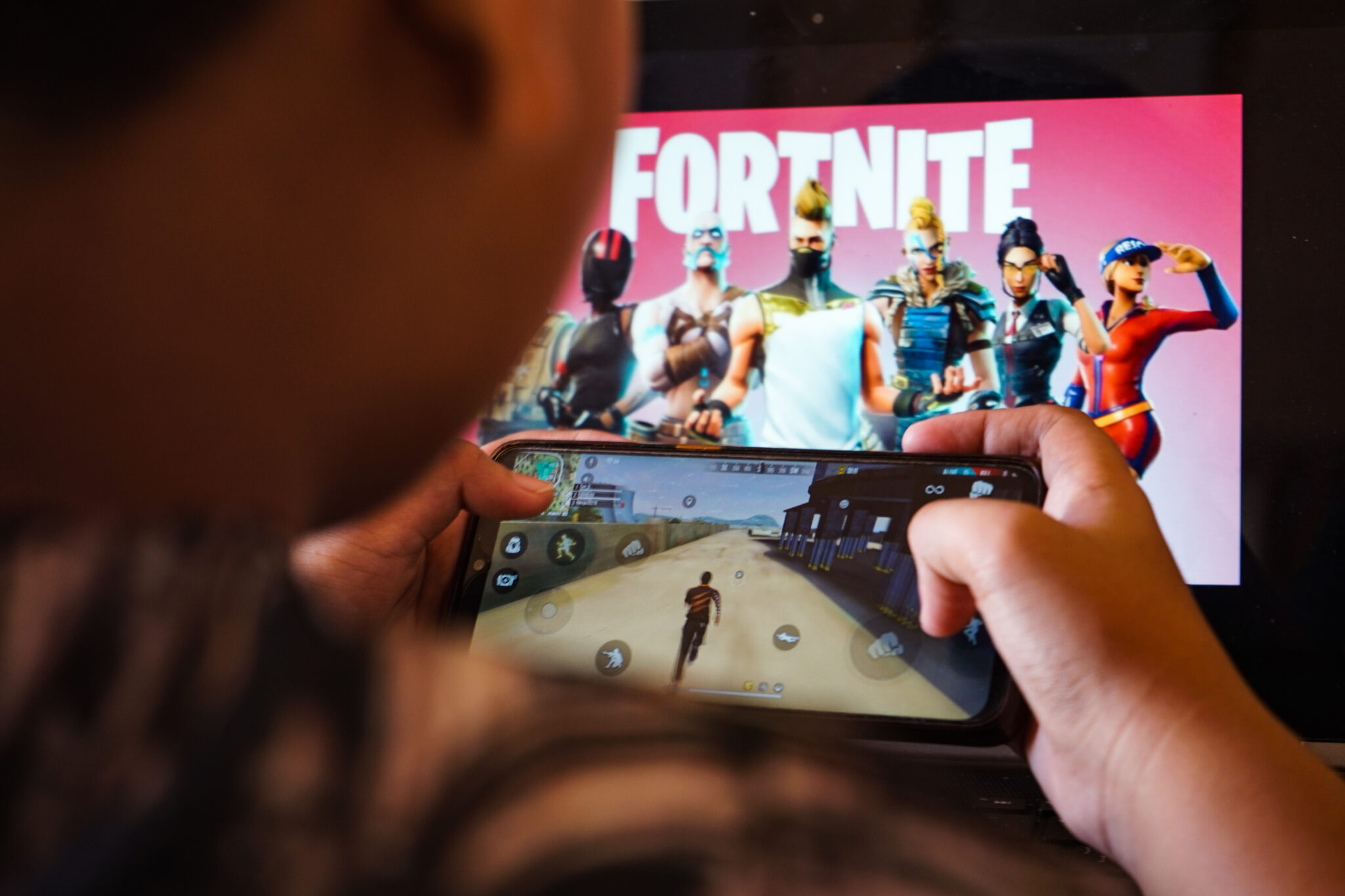 A child plays a game on a cellphone with an image of the Fortnite game on the computer screen in the background.