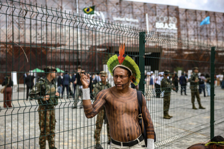 An Amazonian Indigenous man stands outside the fenced perimeter of the COP30 venue in Belém, with military police visible inside the grounds as delegates move through the conference area.