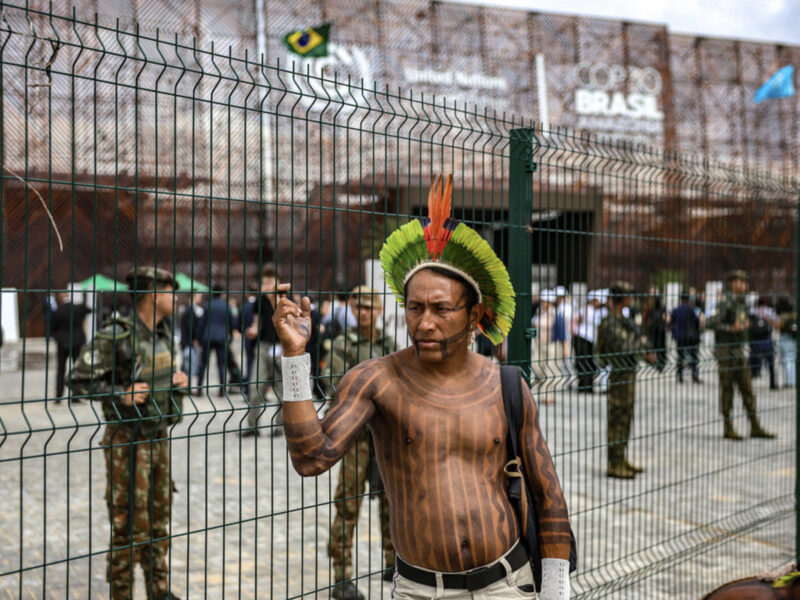 An Amazonian Indigenous man stands outside the fenced perimeter of the COP30 venue in Belém, with military police visible inside the grounds as delegates move through the conference area.