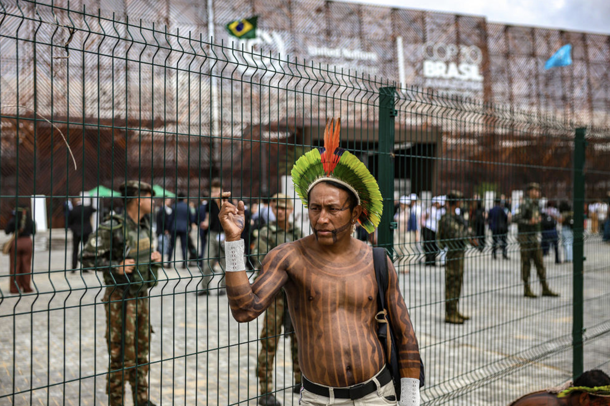 An Amazonian Indigenous man stands outside the fenced perimeter of the COP30 venue in Belém, with military police visible inside the grounds as delegates move through the conference area.